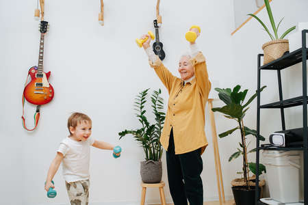 Euphoric granny doing phisical exercises with her grandson, trying to encourage him to stay fit. They are lifting dumbbells overhead.の写真素材