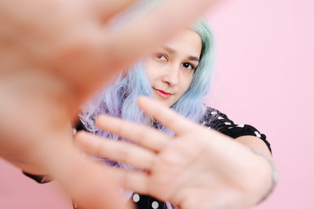 Shy woman in a blue colorful wig and polka dot dress over pink background. She's blocking view on everything except her face.の写真素材