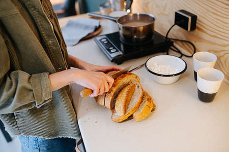 Woman cooking on a high kitchen table, she's cutting bread on a board while rise is boiling on a stove. Cropped, no head. Side view.の写真素材