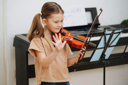 Happy little girl with violin, waving her hand to her grandma in a video chat. The tablet on a music stand. Her beautiful hair in two pony tails.の写真素材