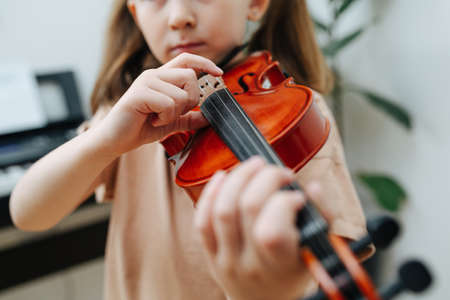 Close up image of a little girl with two pony tails learning to play violin, hitting strings with her finger. She's wearing casual outfit at home.の写真素材