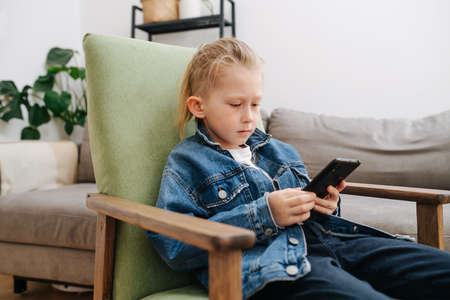 Cool looking little boy sitting in armchair with smartphone in his hands. He is wearing jeans jacket, his thin blond hair brushed back. Low angle.の写真素材