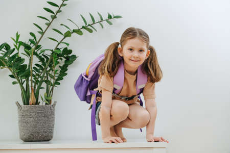 Playful cute little schoolgirl with two pony tails on a white table with purple backpack. She's ready to go school. Over white background.の写真素材
