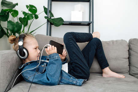 Focused boy in headphones watching his phone on a couch, playing games in the living room. Side view.の写真素材