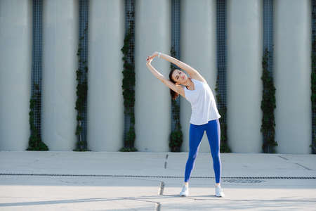 Beautiful woman stretching on a concrete paving, doing side bends. Over support pillars. In blue...