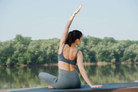 Brunette woman doing yoga outdoors in a beautiful spot on a side of the river. Trees reflecting in the water. On a sunny day under a clear blue sky. She's stretching and twisting in easy pose.の写真素材