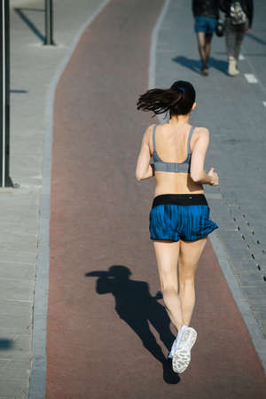 Tall athletic woman running away on the new track outdoors next to a walkway. On a sunny day under a...