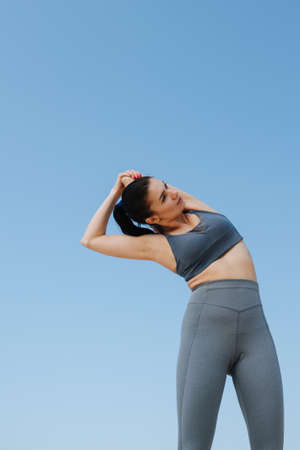 Upward photo of a young brunette woman in gray sportswear doing her stretches. On a sunny day under a clear blue sky.の写真素材