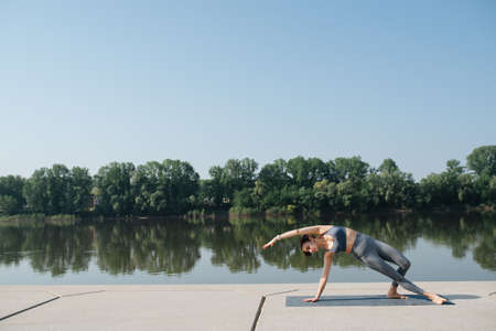 Beautiful graceful brunette doing yoga in a scenic beautiful spot on a riverside. On a sunny day under a clear blue sky. Standing on one hand and legs on the side.の写真素材