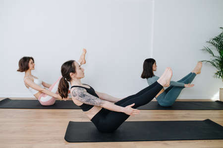 Experienced women in sportswear practicing yoga in a group, holding high boat position in a big studio. Focus on a girl with big shoulder tattoosの写真素材
