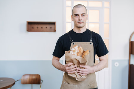 Bold male baker holding paper bag with fresh baguettes. Posing for a photo over a simple indoors setting.の写真素材