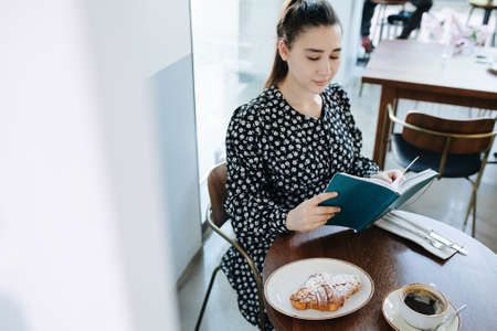 Pretty woman is reading a book in a cafe. Sitting behind a small round table for one person. Wearing black dress with white flower patten.の写真素材