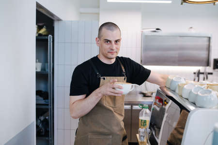 Portrait of a bold male baker posing with coffee cup in hands. Inside a disorderly busy bakery kitchen. Looking at the camera.の写真素材