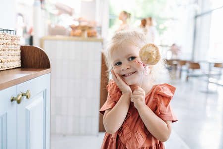 Adorable blonde little girl holding cookie on a stick, smiling at the camera, playing cute.の写真素材
