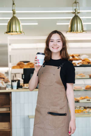 Pretty charming baker girl posing with a plastic coffee cup inside her shop. She's wearing apron, looking at the camera. Over shelves, full of pastry.の写真素材