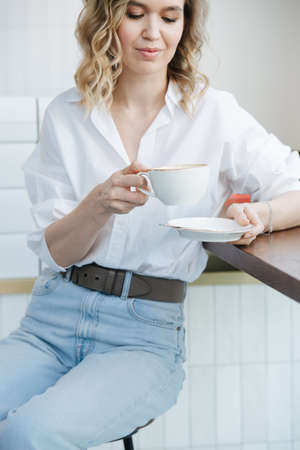 Lonely blond woman sitting on a high stool in a cafel, looking down at a cup of coffee. She's wearing blue dress shirt and light jeans.の写真素材