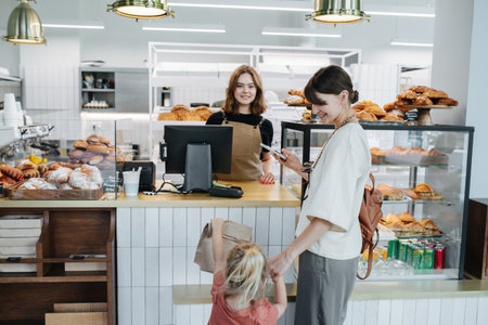 Young woman looking at her happy daughter, holding pack with pastry goods. In a bakery shop. Girl is very lively, showing bag to everyone. Long shot.の写真素材