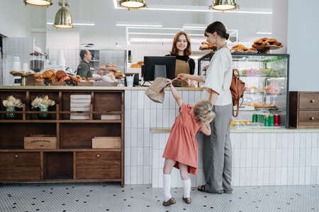 Woman paying for pastry in a bakery shop. Holding daughter by hand. Girl is very restless and impatient. Long shot.の写真素材