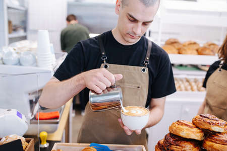 Working male baker putting cream in coffee cup from a metal jug. Inside a bakery kitchen.の写真素材