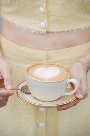 Cream heart in a cup of coffee. In a hands of a woman in a yellow dress. Beautifully presenting it.の写真素材