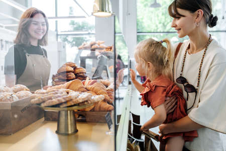 Little girl picking herself some croissants, pointing with index finger, while mother holding her in hands. Cashier smiling at them. Side view.の写真素材