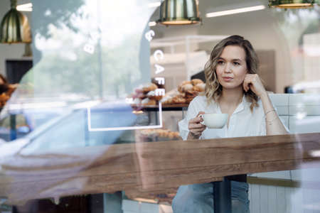Peaceful blond woman enjoying her coffee in a cafe, sitting next to a window, looking at a distance. She's wearing blue dress shirt and light jeans.の写真素材