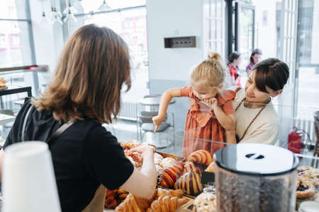 Hungry little girl picking herself some croissants, pointing with index finger, while mother holding her in hands. Cashier helping her choose.の写真素材