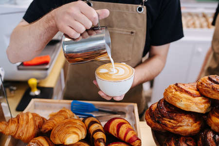 Cropped image of a male baker putting cream in coffee cup from a metal jug. Inside a bakery kitchen next to a pile of pastry.の写真素材
