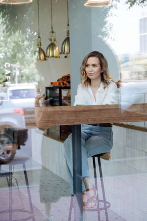 Thoughtful blond woman sitting on a high stool in a cafe next to a window. She's wearing blue dress shirt and light jeans, looking at a distance.の写真素材