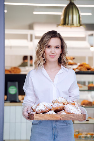 Bokeh portrait of a blond woman posing with a tray full of pastry in a doorstep of her shop. She's wearing dress shirt and light jeans, looking at the camera.の写真素材