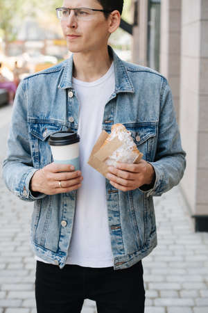 Man in a jeans jacket satisfied about his purchase in a bakery. Holding coffee and a croissant, wrapped in a paper.の写真素材