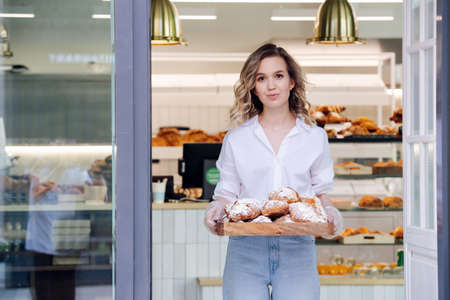 Tight lipped blond woman posing with a tray full of pastry in a doorstep of her shop. She's wearing dress shirt and light jeans, looking at the camera.の写真素材