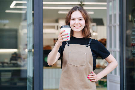 Cheeky smiling baker girl posing with a plastic coffee cup in front of her shop. She's wearing apron, looking at the camera.の写真素材