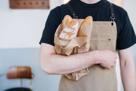 Paper bag with fresh baguettes held by a male baker in beige apron. Posing for a photo over a simple indoors setting.の写真素材