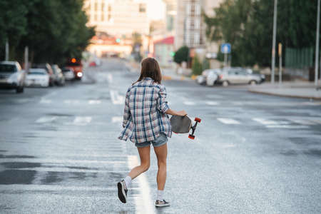 Woman walking across wet empty city road with skateboard in hand early in the morning. She's wearing checkered shirt and mini jeans shorts. Facing away.の写真素材