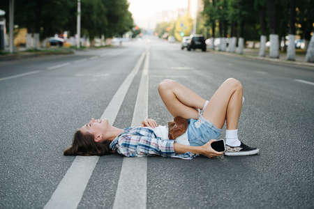 Crazy lighthearted young woman lying on an asphalt road, Having a laugh about situatuon. Side view. She's wearing checkered shirt and mini shorts.の写真素材