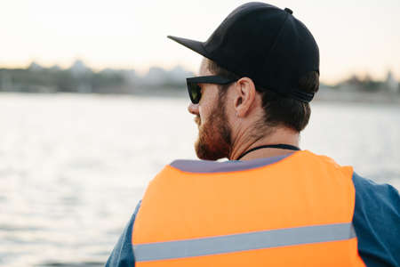 Bearded man in a life vest sitting in front of a large body of water. He's wearing black sun cap and sunglasses. Looking to the side. From behind.の写真素材