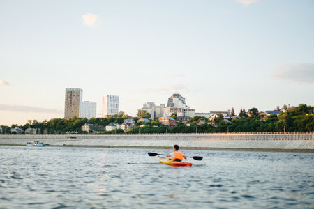 Longshot of a man kayaking on a big wide river next to a reinforced concrete riverbank. Wooden and high storey buildings in a distance.の写真素材