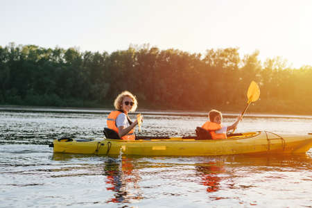 Smiling mother and daughter in orange life jackets kayaking on a yellow boat on a big wide river. Side view. They are looking at the camera.の写真素材