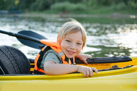 Enthusiastic blond toddler boy enjoying his ride on yellow kayak on a big wide river. Vearing life vest. Smiling at the camera.の写真素材