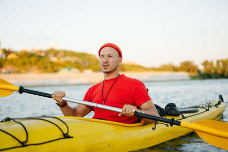 Bold man in orange watch cap and shirt riding on yellow kayak on a big wide river. Reinforsed shoreline, trees and some houses in a distance.の写真素材