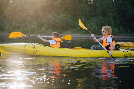 Mother and daughter in orange life jackets kayaking on a yellow boat on a big wide river. Side view. Sun relecting beautifully in a green water.の写真素材