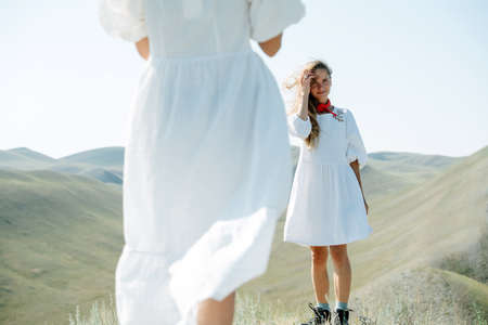 Two women in simple thin white mini dresses standing on a peak amidst beautiful hills. Standing one in front of another,looking at each other. From behind one of them. Middle part of the body.の写真素材