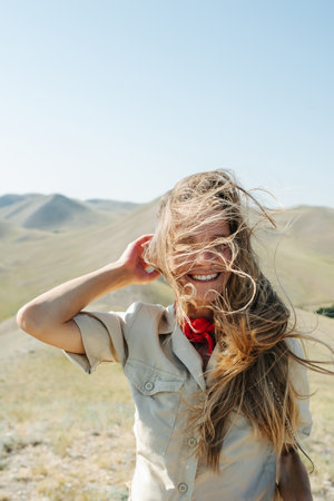 Happy woman with windy messed up hair posing amidst a dry field. Stunning view on beautiful hills...