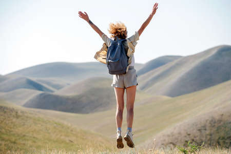 Euphoric woman jumping on the spot amidst a beautiful hills scenery. Throwing hands in the air. From behind.の写真素材