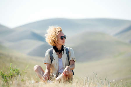 Happy woman with curly messed up hair sitting amidst a dry field, taking in stunning view on beautiful hills stretching all across.の写真素材