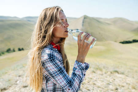 Thirsty woman drinking water. Exhausted from the hike. Surrounded by grassy beautiful hills, stretching far across.の写真素材