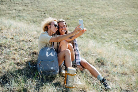 Best girl friends sitting on a dry grass slope, taking selfie, smiling. Sitting closely together.