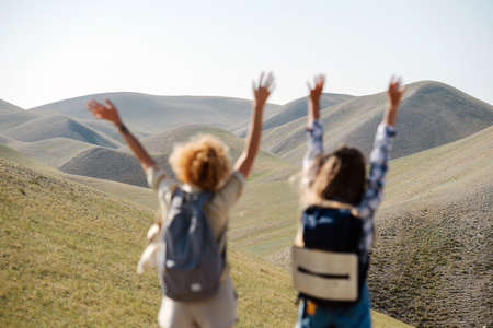 Exhilarated girl friends jumping on the spot amidst a beautiful hills scenery. Throwing hands in the air. From behind.の写真素材