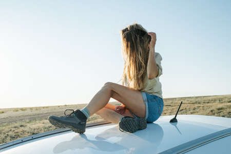 Restful woman sitting on a car roof, enjoying wind and views on scenic steppe plain. Her hair is...